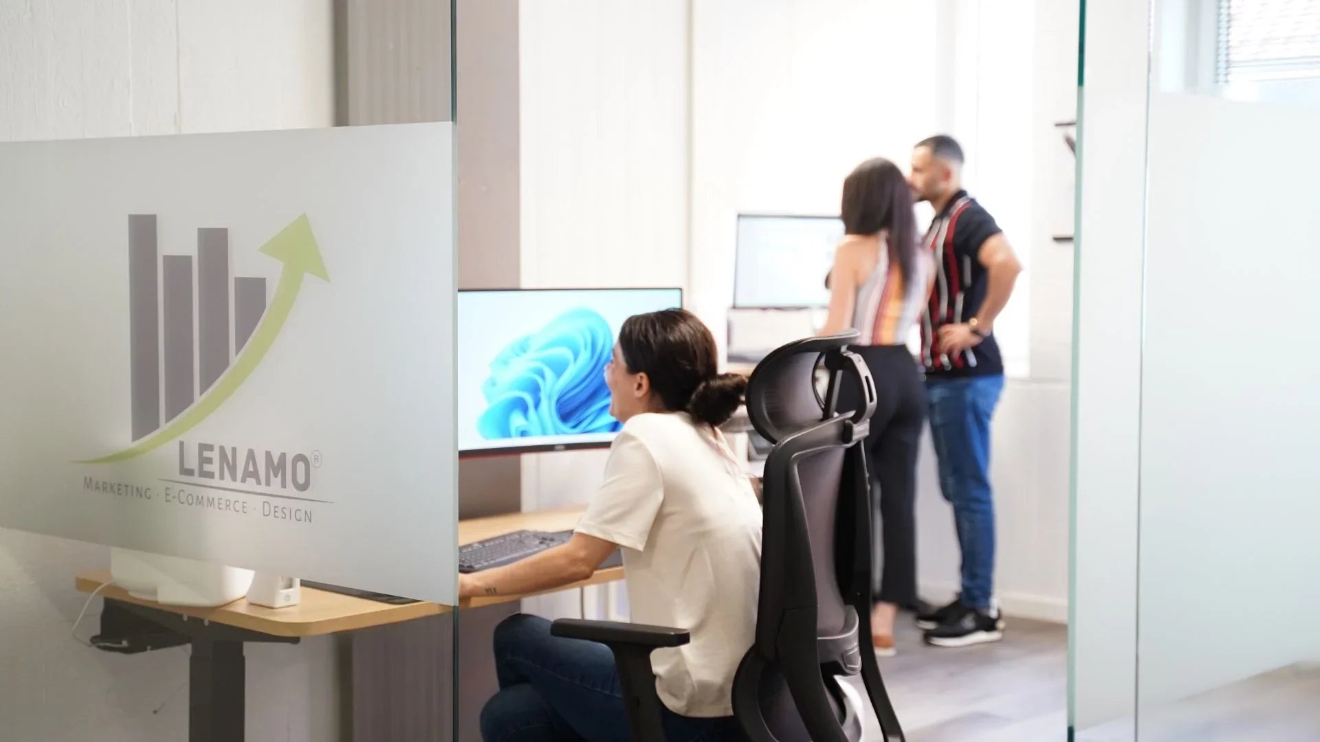 Eine Dame sitzt an ihrem ergonomischen Arbeitsplatz und zwei Personen stehen zusammen an einem ergonomischen Arbeitsplatz