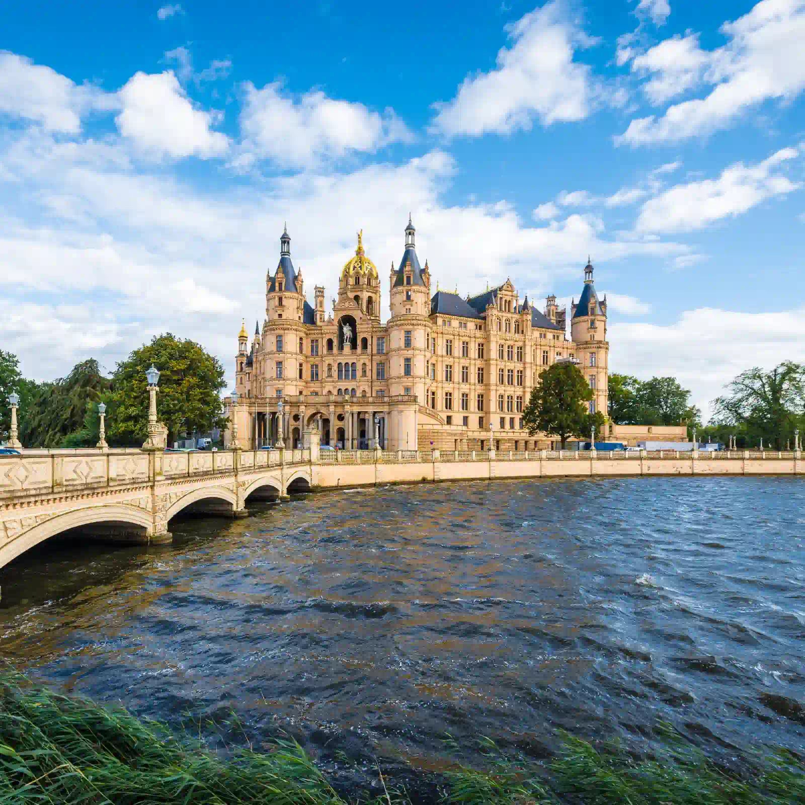 Das Schloss in Schwerin bei leicht bewölktem Tag. Grüne Bäume und leichte Wellen im Wasser.