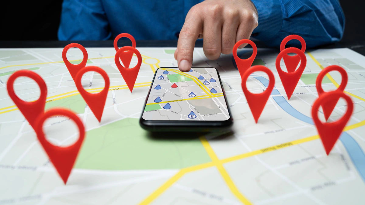 Smartphone liegt auf einer Landkarte mit mehreren roten Pins als Markierung verschiedener Standorte. Eine männliche Hand bedient das Smartphone mit einem Finger.